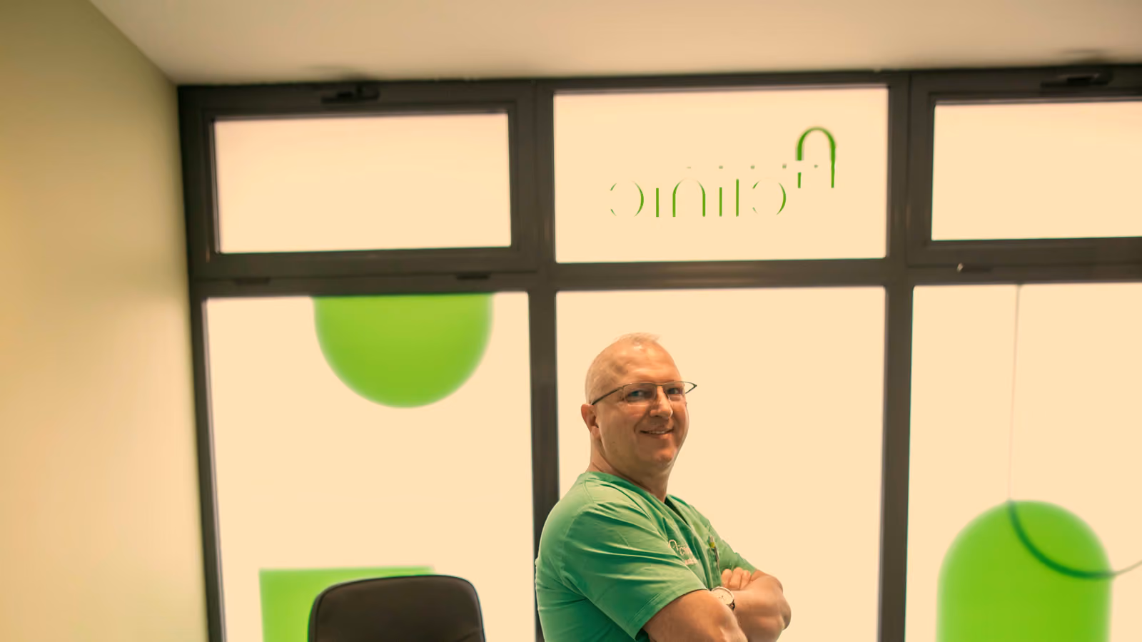 Medic în halat verde A Clinic, în cabinetul clinicii, cu geamurile și logoul clinicii în spate