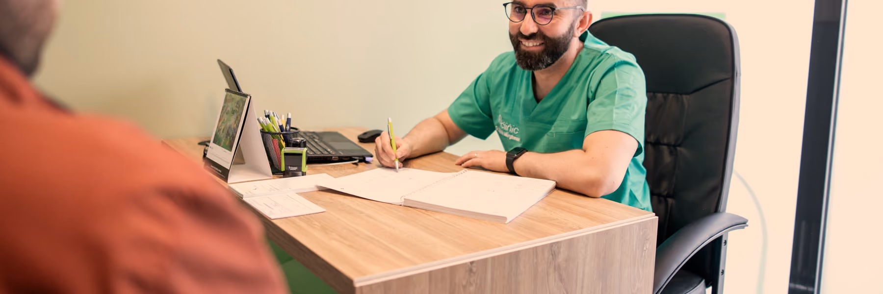 Medic în halat verde A Clinic, la birou, notând pe o fișă în timpul unei consultații, cu pacientul în prim-plan