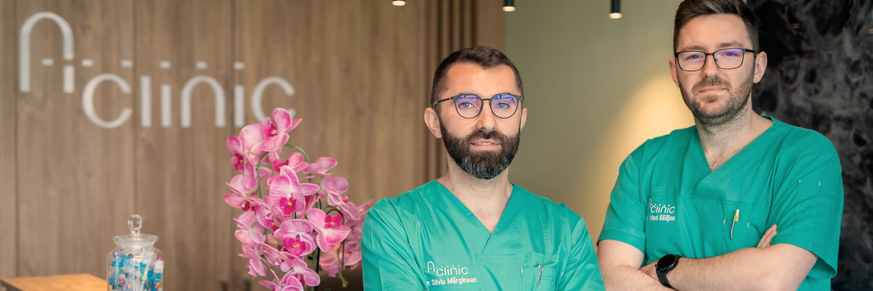 Doi medici A Clinic la recepția clinicii, în fața peretelui de lemn cu logoul și a orhideei