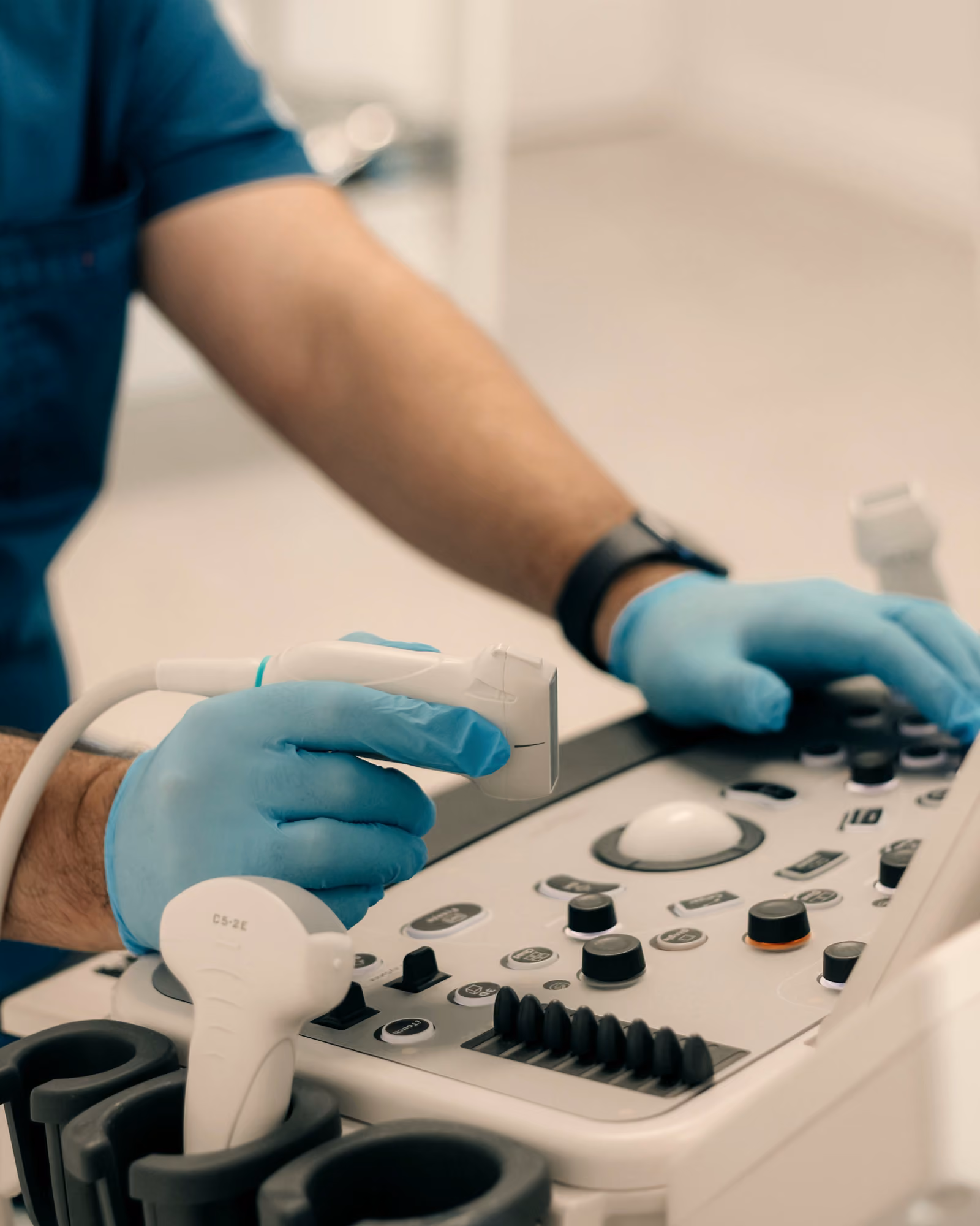 Mâini cu mănuși medicale pe consola unui ecograf, una dintre ele ținând transductorul