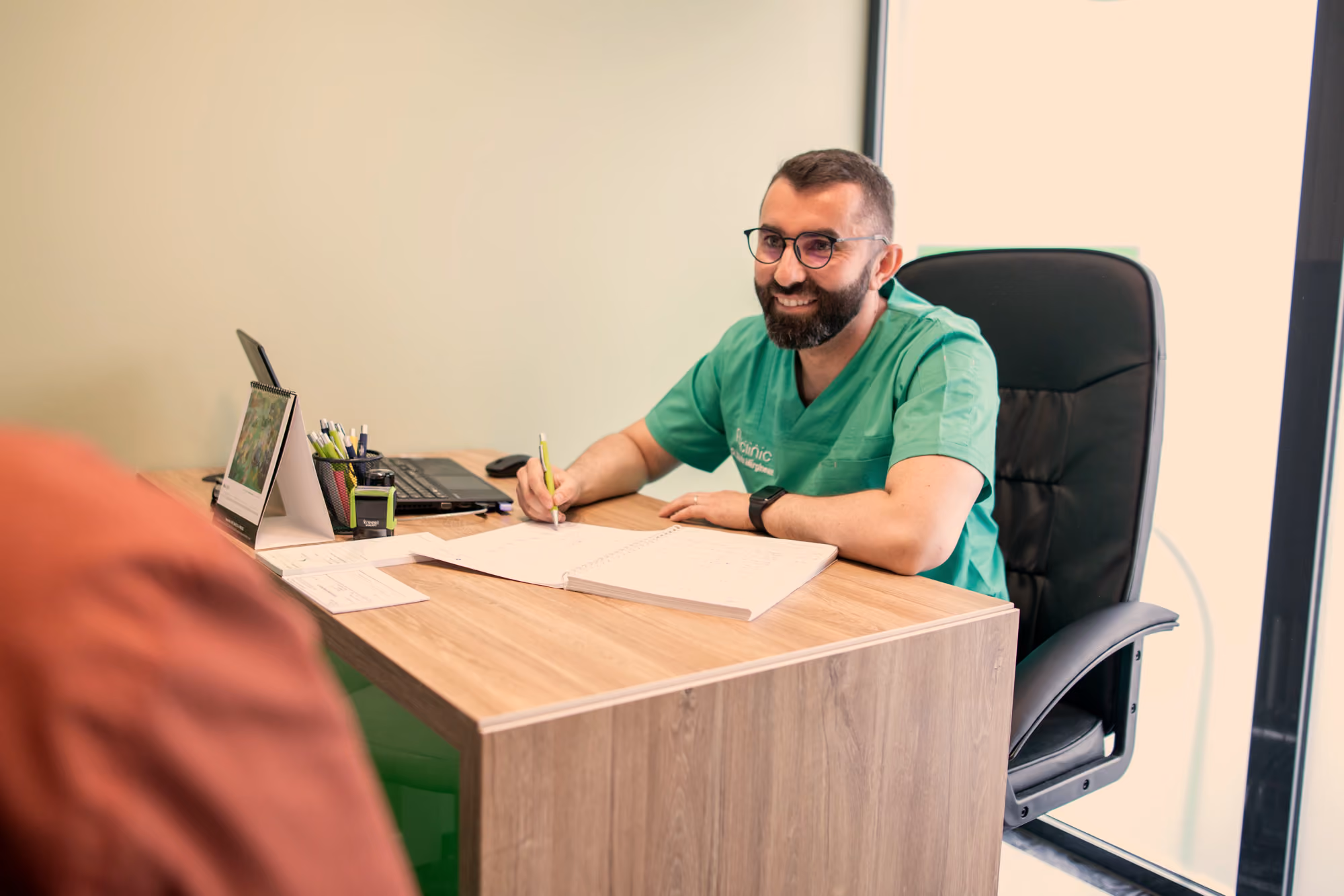 Medic A Clinic în halat verde, la biroul din cabinetul de consultații, în discuție cu un pacient