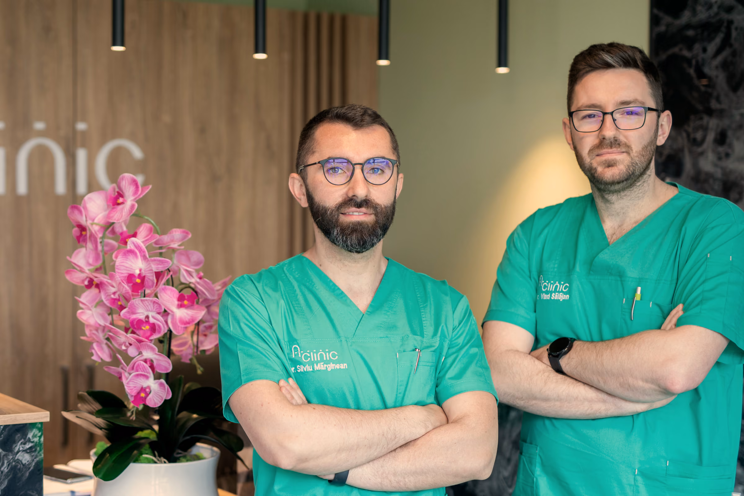 Dr. Silviu Mărginean și dr. Vlad Sălăjan, în halate verzi A Clinic, cu brațele încrucișate, la recepția clinicii