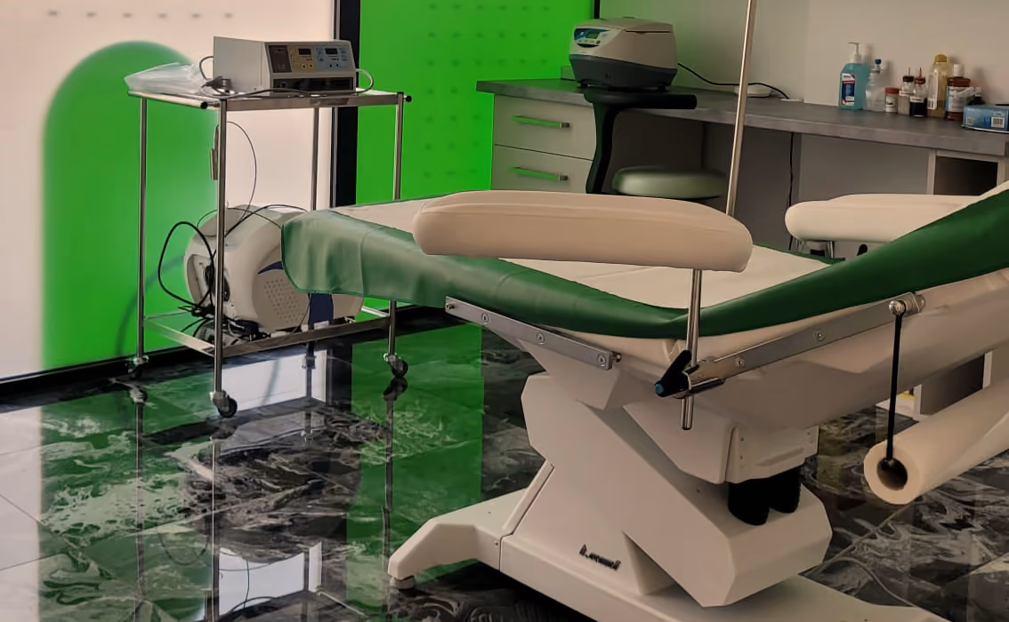 Sală de tratament A Clinic, cu masa de procedură verde cu alb și căruciorul cu aparatură, lângă peretele de sticlă verde