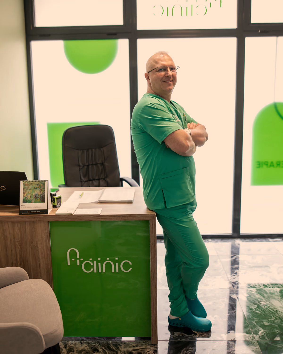 Dr. Claudiu Mare, în halat verde, sprijinit de biroul din cabinetul A Clinic